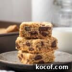 Three chocolate chip cookie bars stacked on a plate with a glass of milk in the background.