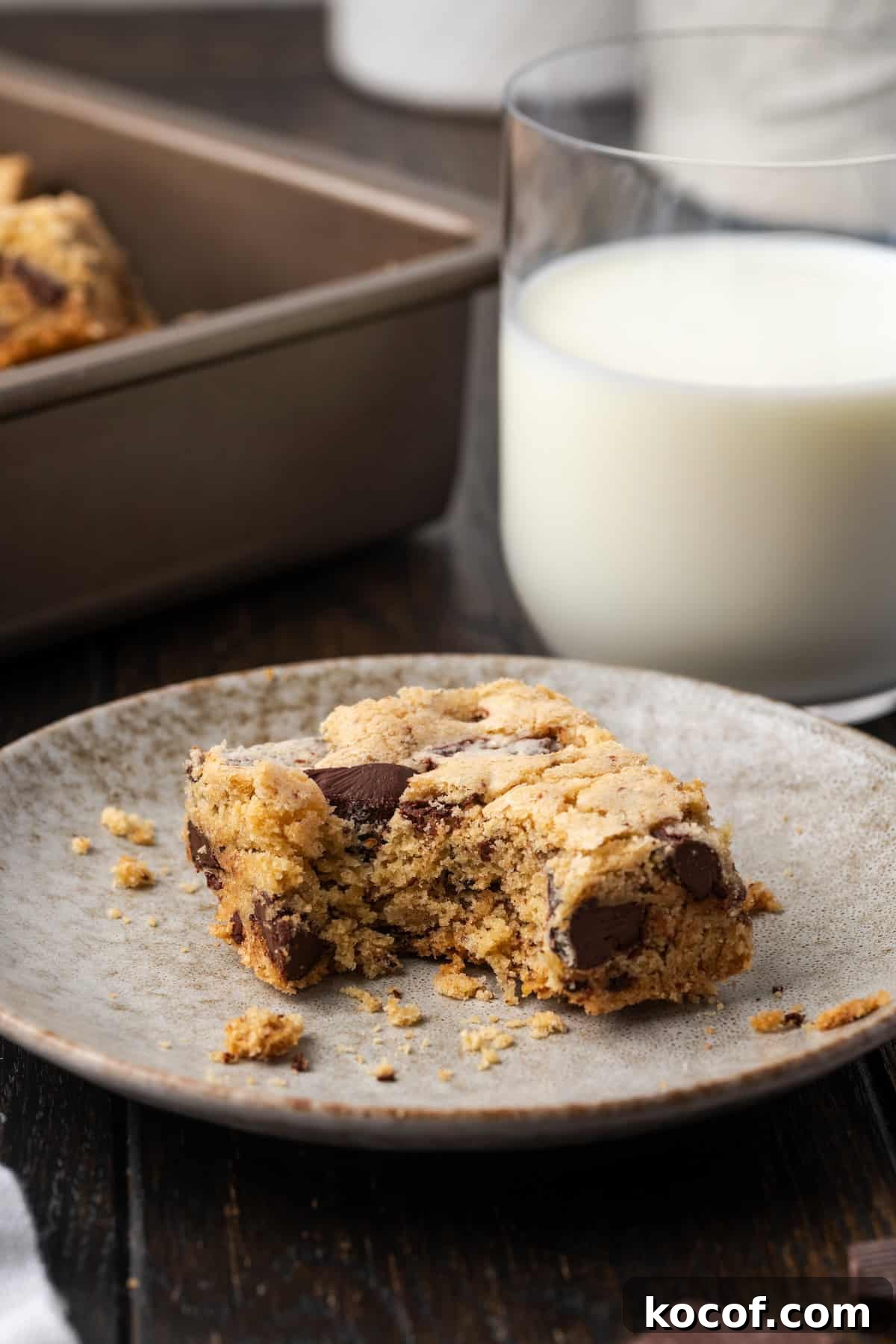 Buttery, chewy chocolate chip cookie bars are easy to make and perfect for baking and slicing into squares, no chilling or rolling required! A chocolate chip cookie bar with a bite missing on a plate, with a glass of milk on the background.