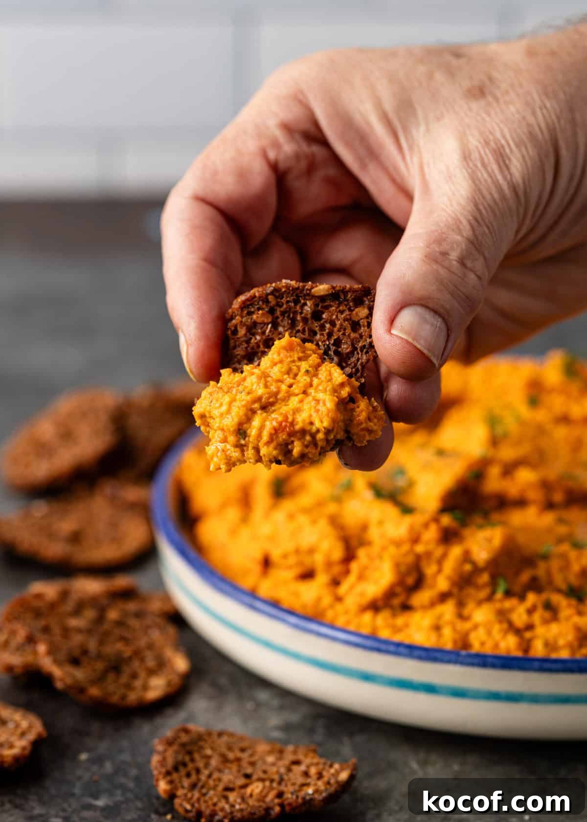 Roasted Carrot and Goat Cheese Dip Recipe for Entertaining 9 closeup side view: hand holding cracker with goat cheese dip over top of bowl of carrot and goat cheese dip and crackers on counter