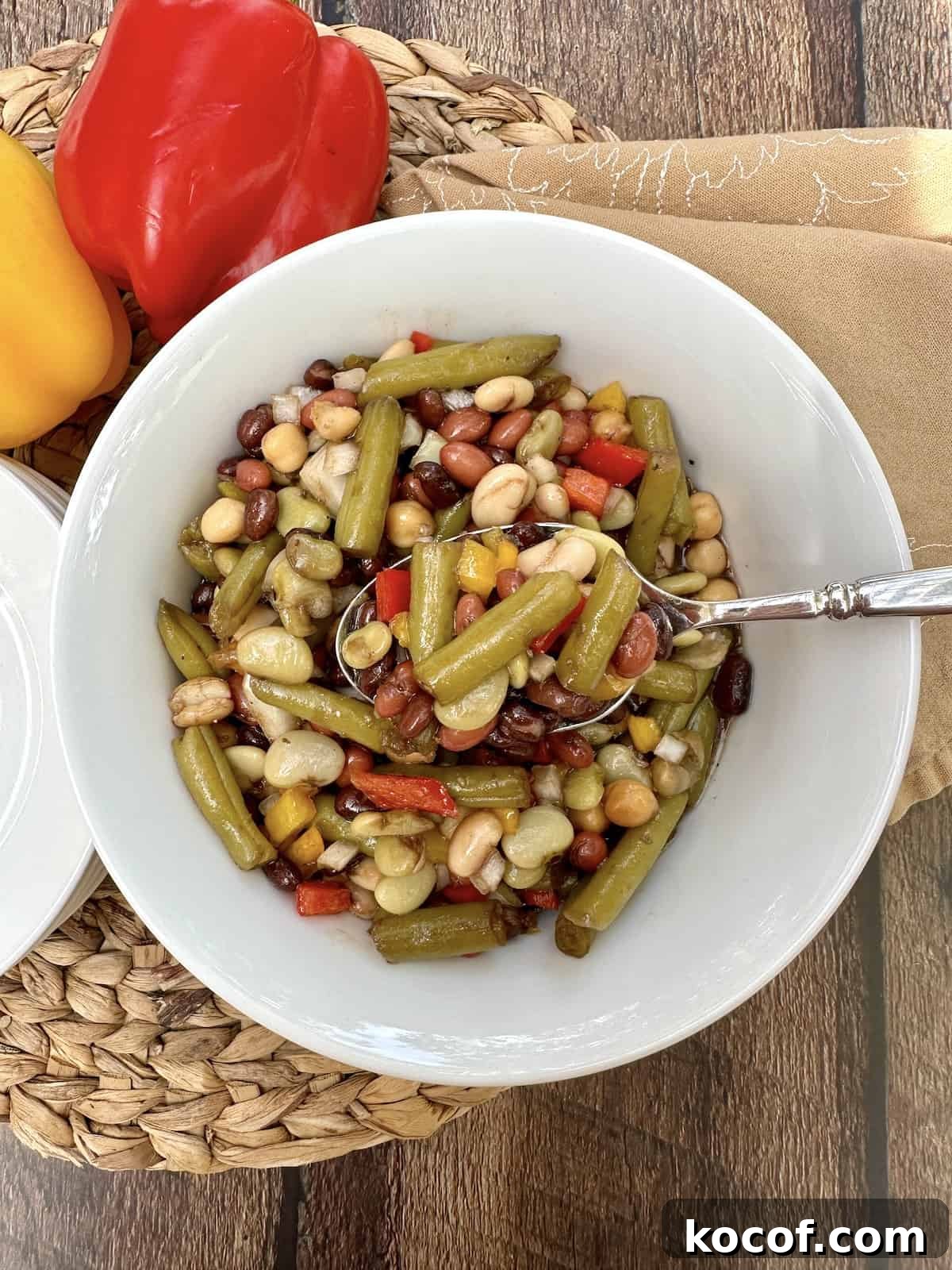 A serving bowl of cold green bean salad with a spoon, on a place mat with two peppers off to the side.
