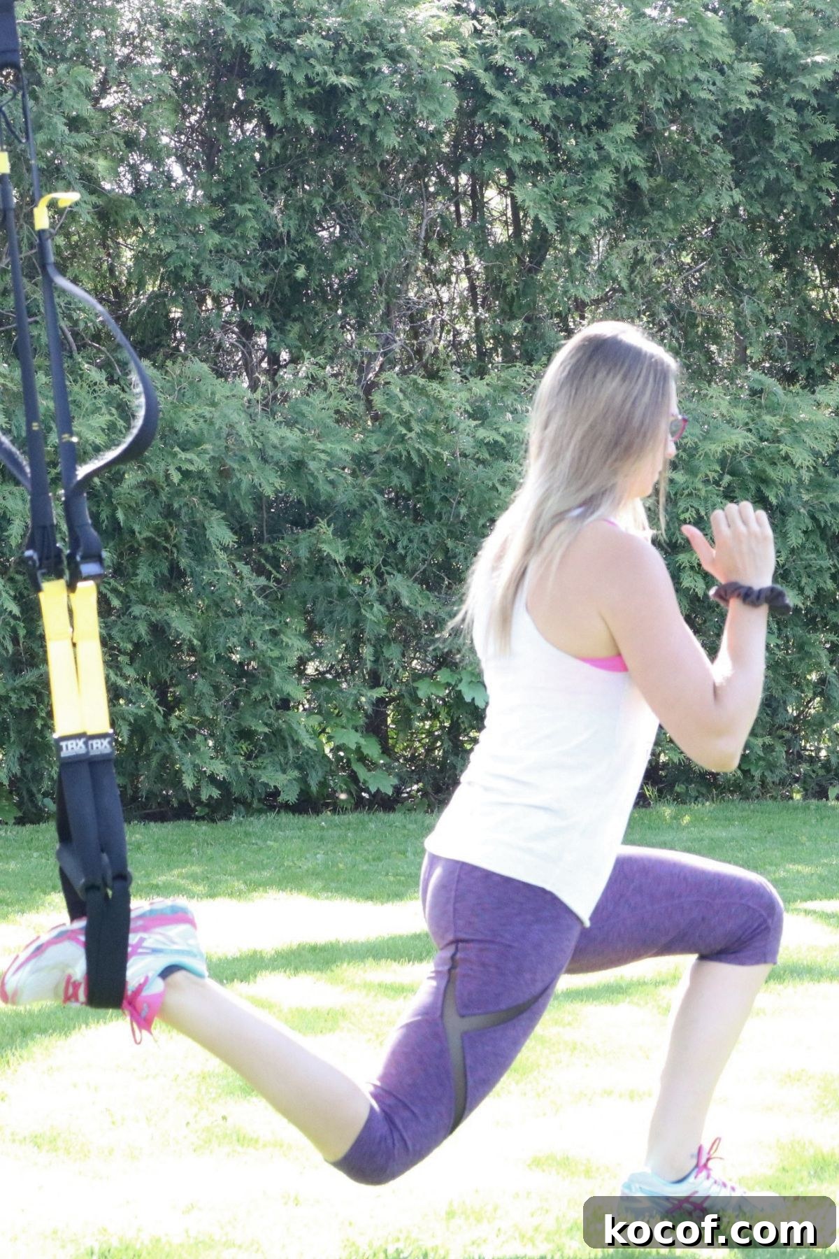 Woman in white shirt and purple pants performing a TRX lunge.