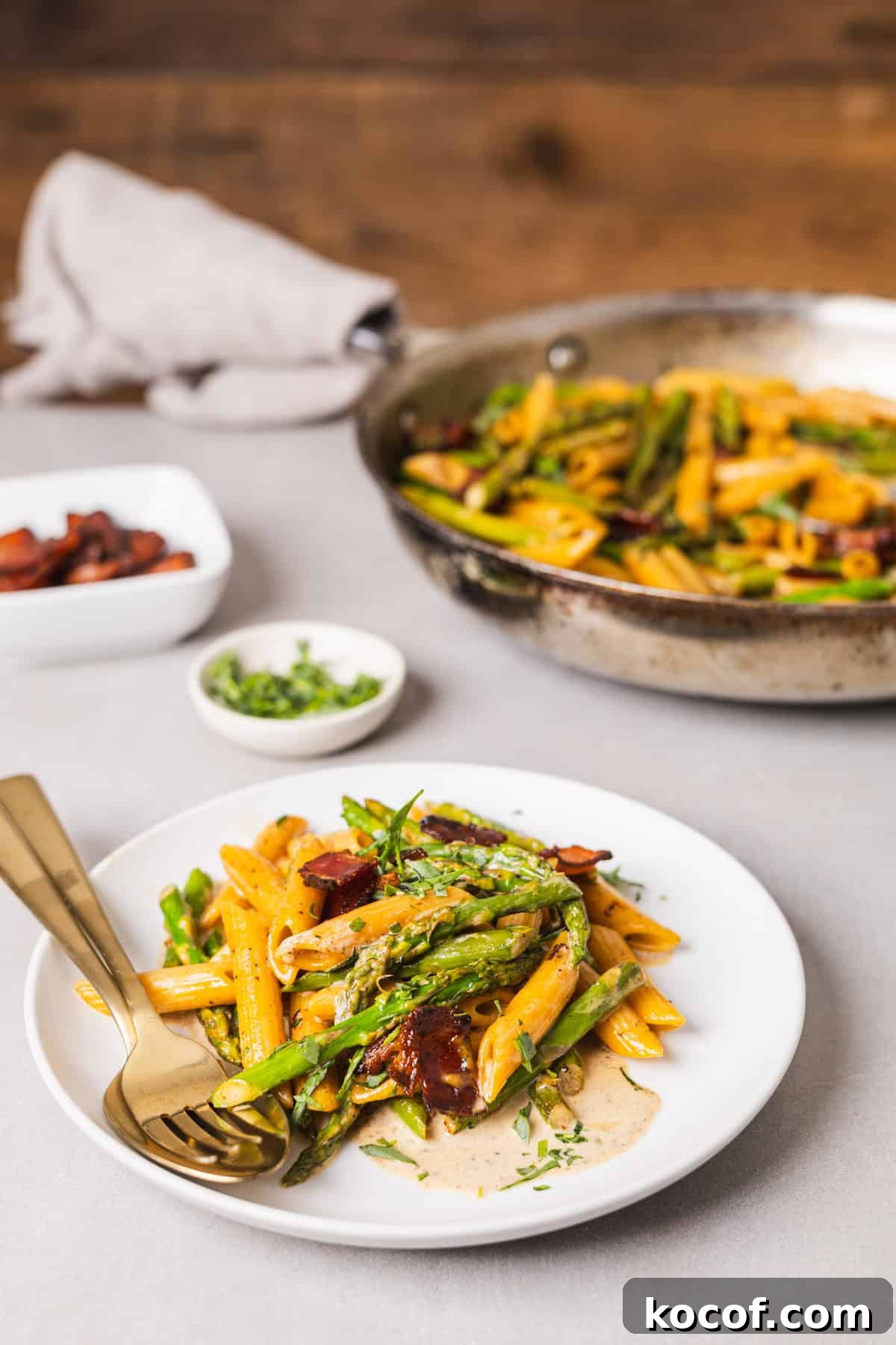 Creamy Bacon and Asparagus Pasta Recipe for Spring Meals 8 A white plate with bacon and asparagus pasta with a skillet with more pasta in the background.