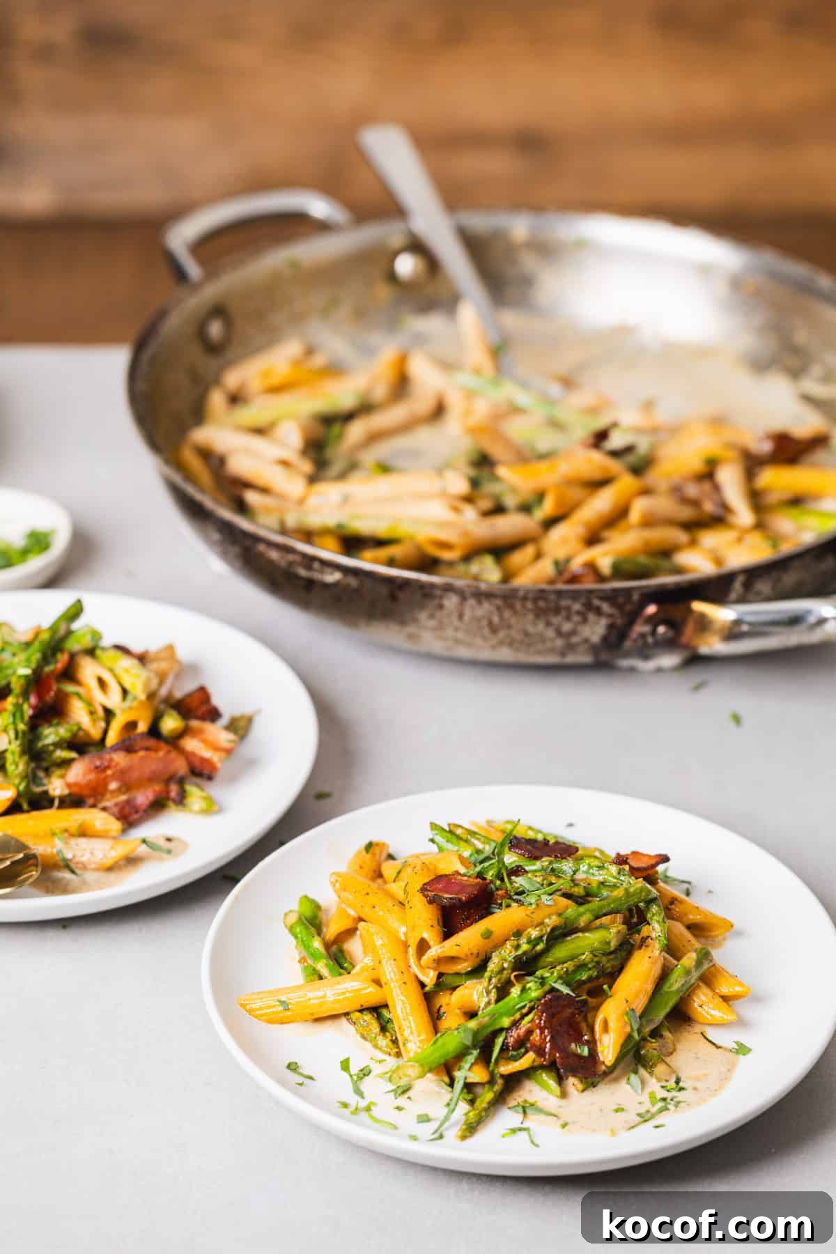 Creamy Bacon and Asparagus Pasta Recipe for Spring Meals 9 Two small plates with bacon and asparagus pasta on a gray table and a skillet with more pasta in the background.