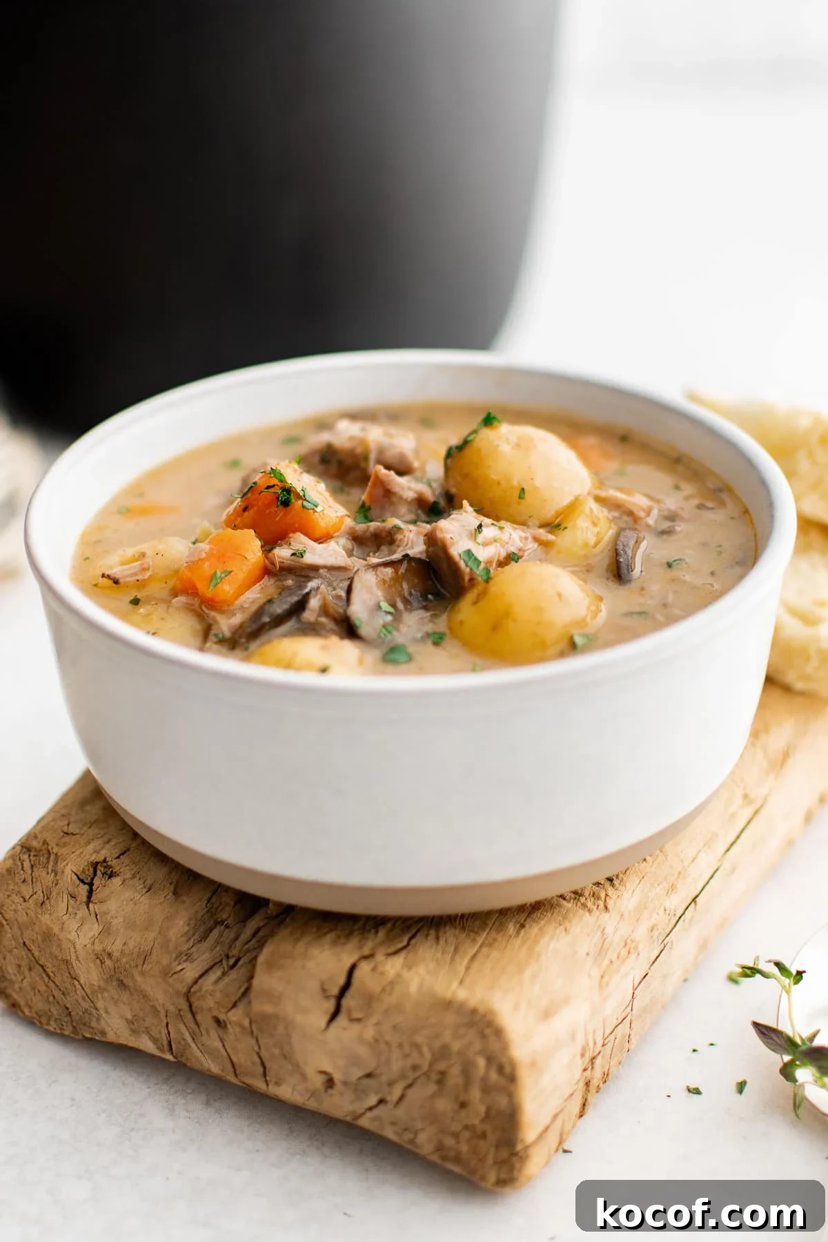 Side view of a white ceramic bowl filled with creamy pork stew on a rustic wooden board, with pork chunks, baby potatoes, carrots, and mushrooms visible in the thick gravy and topped with chopped green herbs.