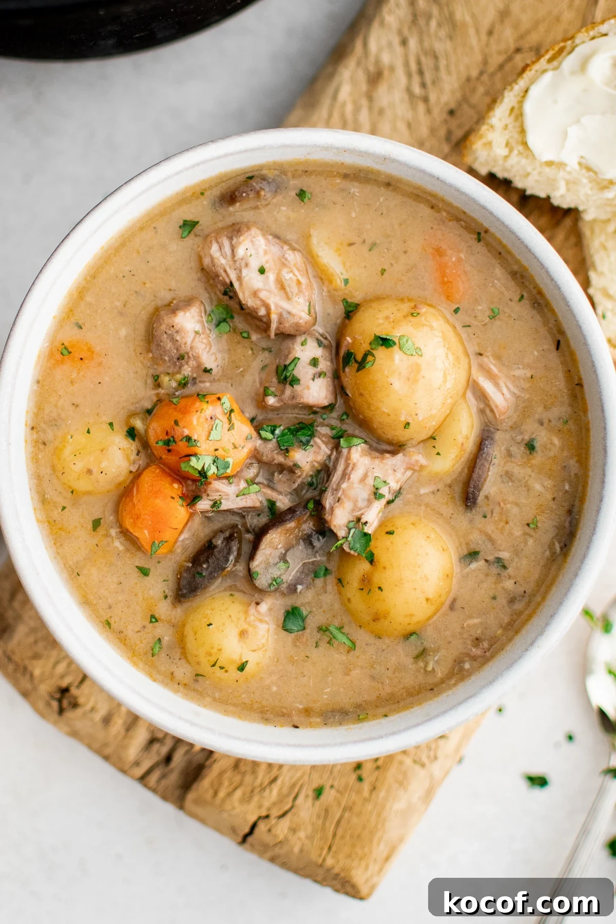 Overhead view of a white bowl of creamy pork stew on a wooden board, filled with pork chunks, baby potatoes, carrots, and mushrooms in a thick beige gravy and sprinkled with chopped green herbs, with a piece of bread spread with butter visible at the top right.