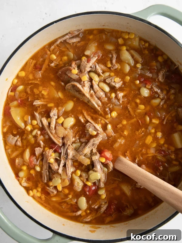 Large Dutch oven filled with a stew made with shredded pork, frozen lima beans, corn, tomatoes, potatoes, onions, and bbq sauce.