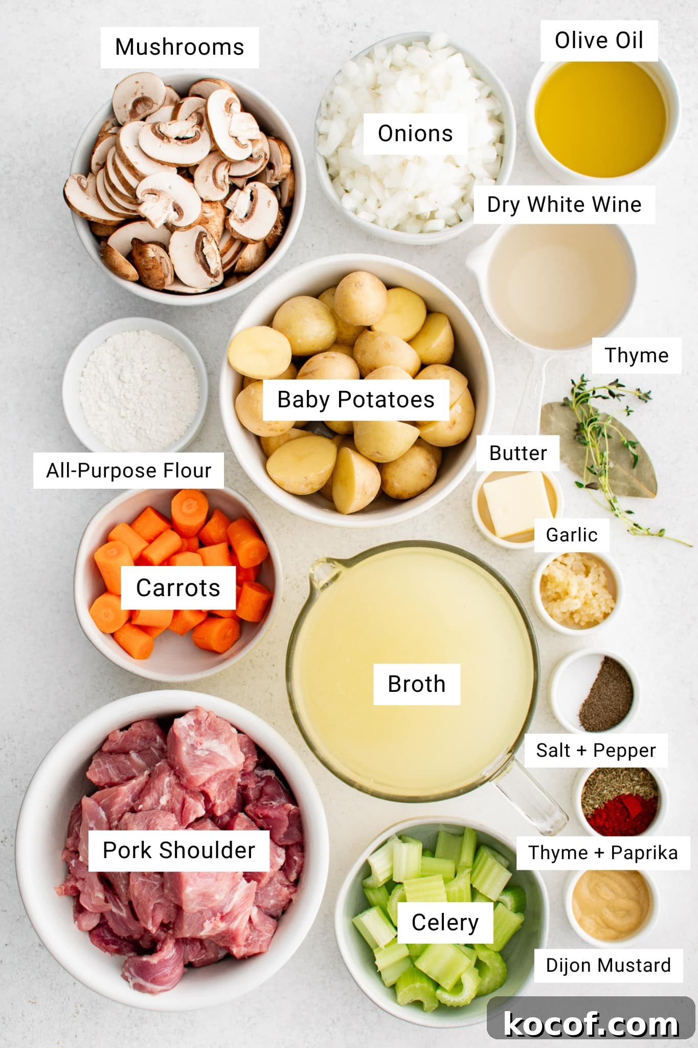 Overhead ingredient photo on a light gray surface with labeled white text boxes showing bowls and ingredients for stovetop pork stew, including sliced mushrooms, chopped onions, olive oil, dry white wine, all-purpose flour, carrots, pork shoulder pieces, celery, broth, garlic, butter, thyme, salt and pepper, paprika, Dijon mustard, and baby potatoes.