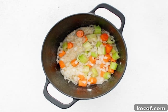 Overhead view of chopped onions, sliced carrots, and chunky celery pieces in a black Dutch oven beginning to soften.