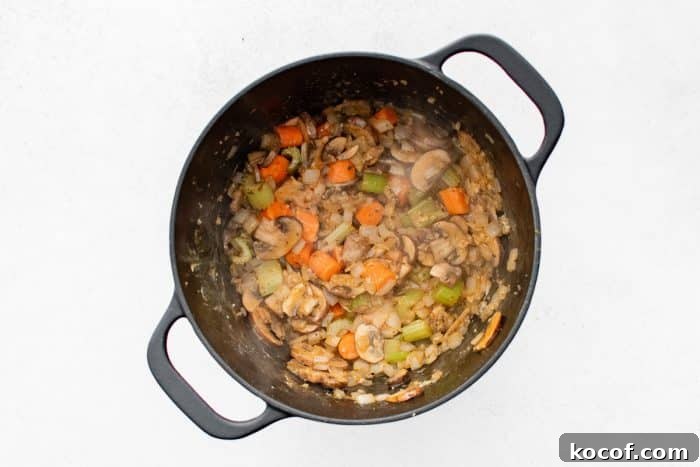Softened onions, carrots, celery, and mushrooms cooking in a black Dutch oven, lightly browned and steamy.