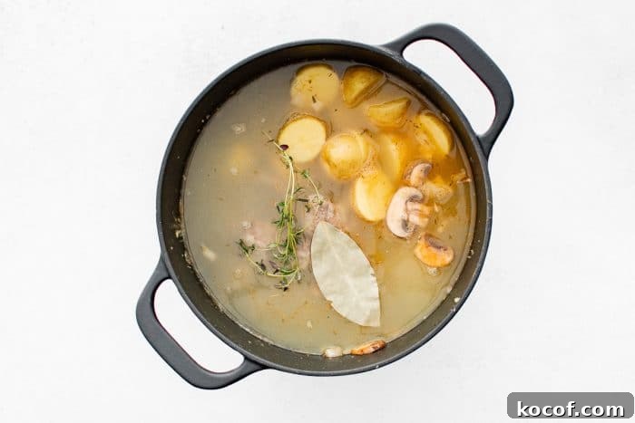 Overhead view of a black Dutch oven filled with broth and halved baby potatoes, with a bay leaf and thyme sprigs floating on top.