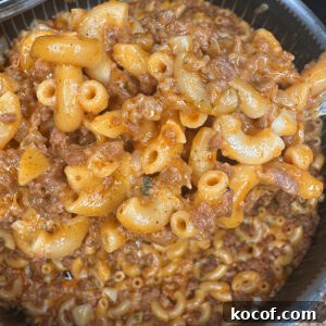 A pot filled with cooked macaroni, ground beef, and a rich tomato-based sauce—reminiscent of classic Hamburger Helper—rests on a woven mat. A metal spoon is partially visible, stirring the hearty mixture.
