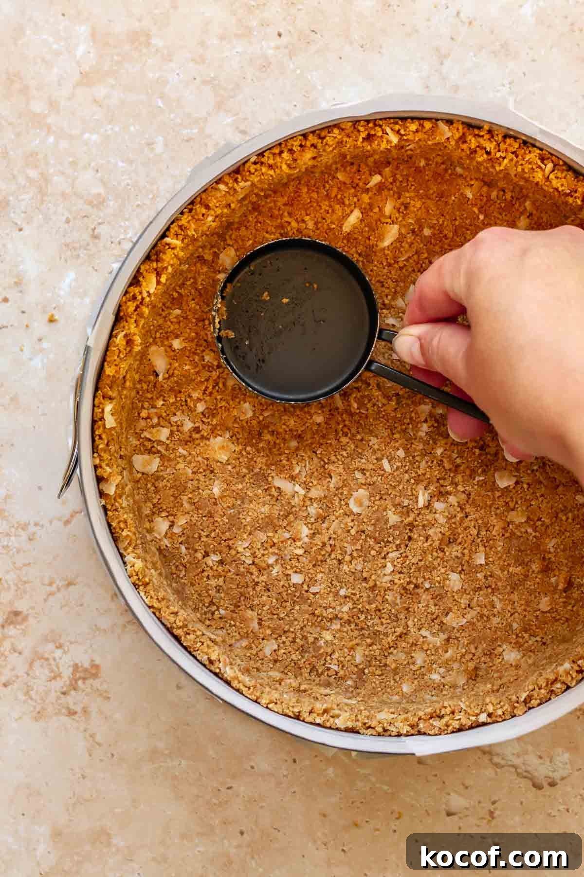 Creamy Ube Cheesecake Recipe: Filipino Purple Yam Dessert 6 A hand uses a measuring cup to press graham cracker crust into a pan.
