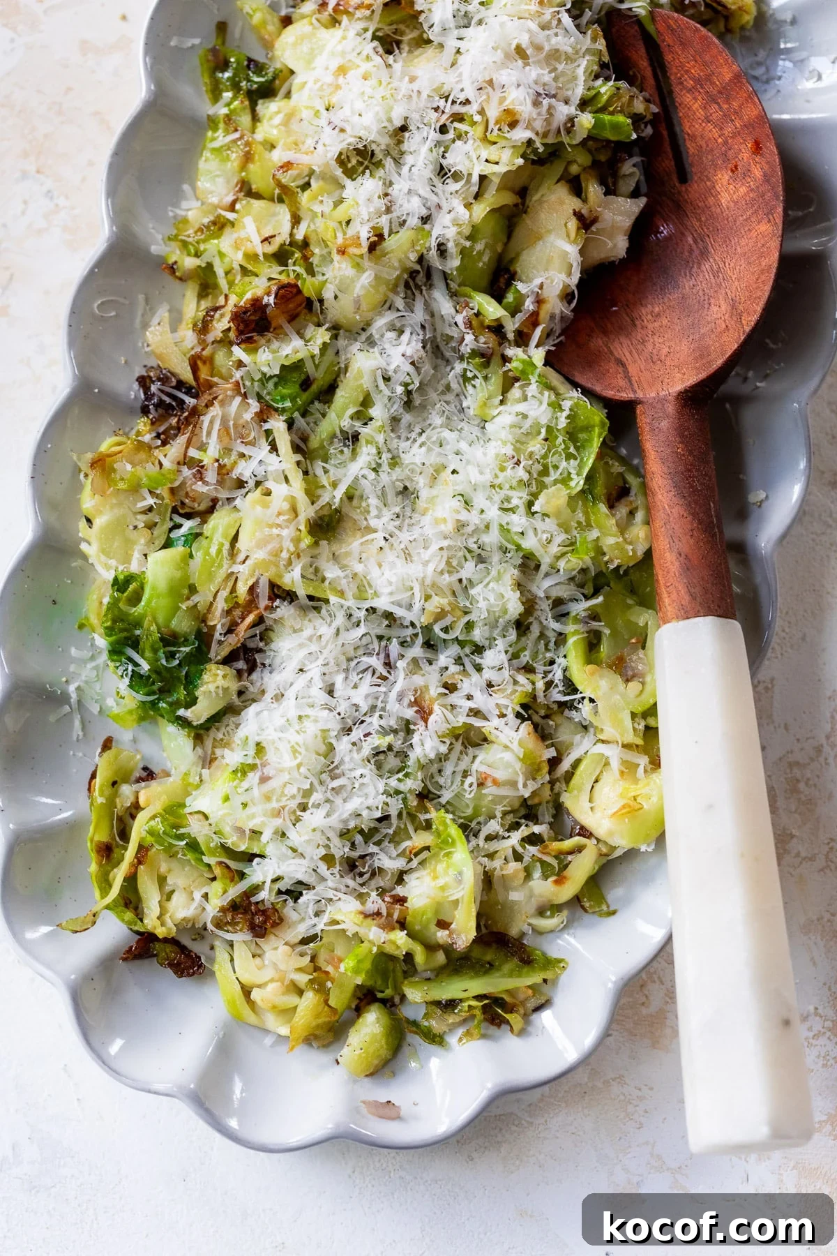 shaved brussels sprouts topped with shredded cheese next to a wooden spoon on a white serving platter.