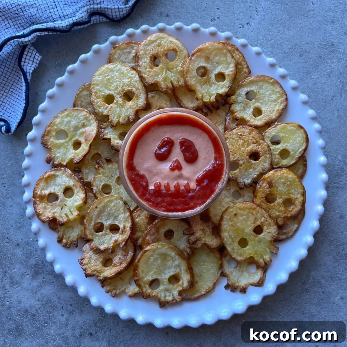 A plate of skull-shaped potato slices arranged around a bowl of dip with a skull face design made from sauce on top.