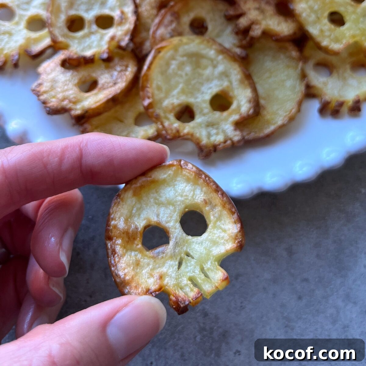 A hand holds a skull-shaped potato chip in front of a plate with more skull-shaped chips on it.