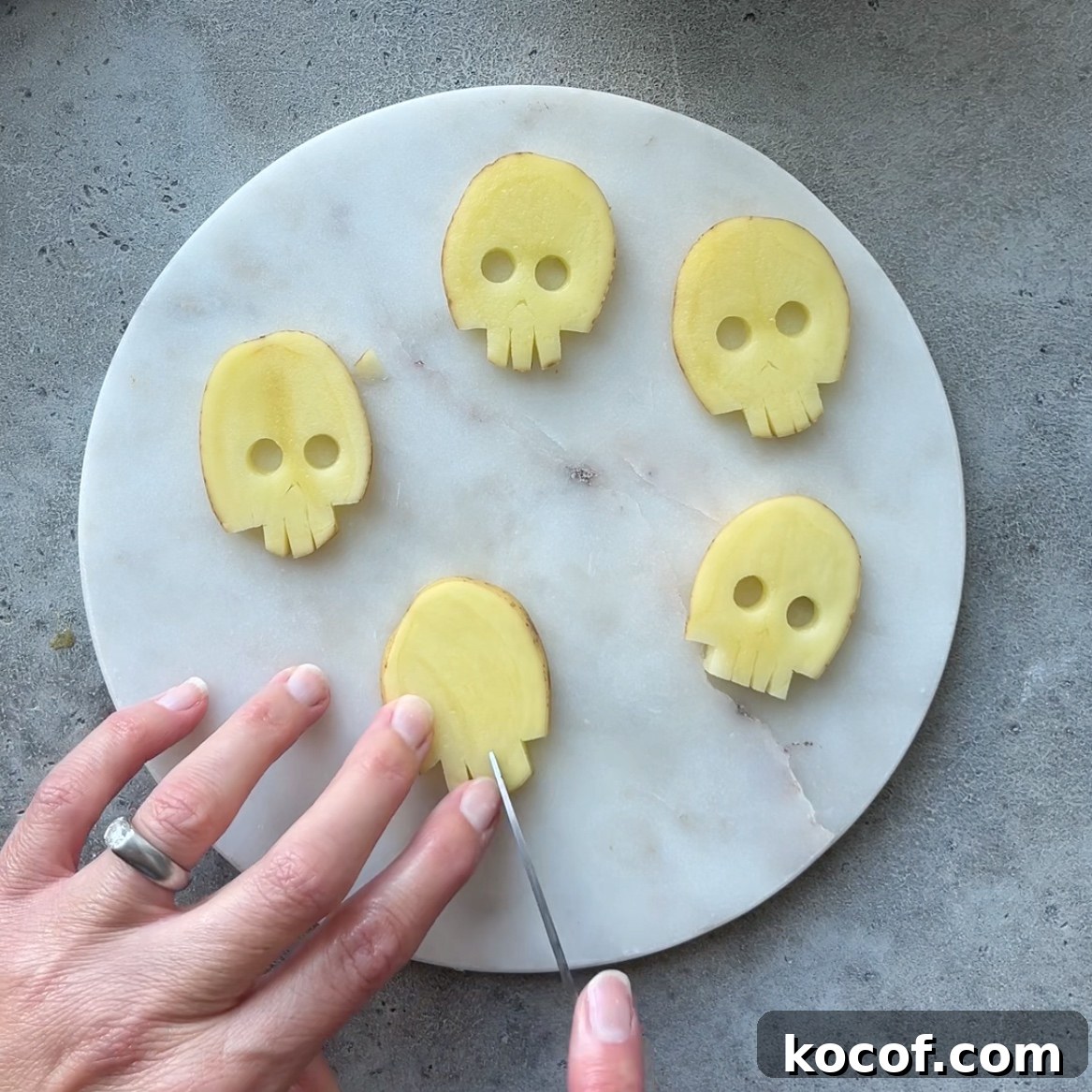 Four thin potato slices cut into skull shapes are arranged on a round marble board. A hand is carving details into another slice.