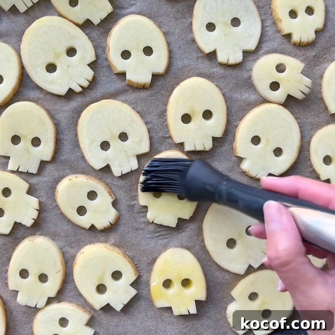 Potato slices cut into skull shapes are arranged on a baking sheet. A hand is brushing oil onto the slices with a basting brush.