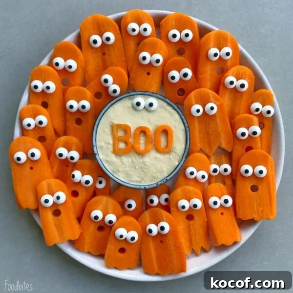 A plate of Halloween vegetables features carrot slices shaped like ghosts with candy eyes, arranged around a bowl of dip with BOO spelled on top.