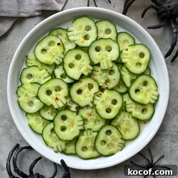 Cucumber slices carved to look like skulls are arranged on a white plate, sprinkled with red pepper flakes for a healthy twist. Plastic black spiders surround these festive bites, making them perfect for Halloween-themed food recipes.