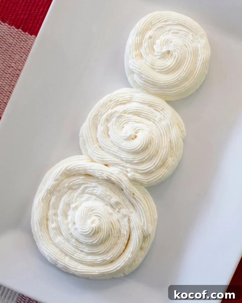 Snowman Cheesecake Dip Recipe for Holiday Parties 3 Three swirls of piped white frosting arranged vertically on a white rectangular plate, resembling a snowman shape. The background includes part of a red and white checkered cloth.