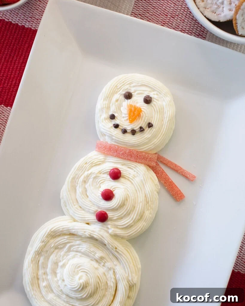 Snowman Cheesecake Dip Recipe for Holiday Parties 4 A snowman-shaped dessert made of swirled white frosting, decorated with chocolate chips for eyes and mouth, an orange candy nose, red candies as buttons, and a red gummy candy scarf, arranged on a white platter.