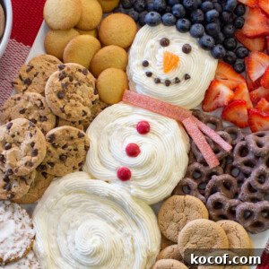 Snowman Cheesecake Dip Recipe for Holiday Parties 6 A festive dessert platter features a snowman made of swirled white frosting, decorated with blueberries, strawberries, candy, and surrounded by various cookies and chocolate-covered pretzels.