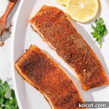 Two baked blackened salmon fillets on a white plate with lemon slices.