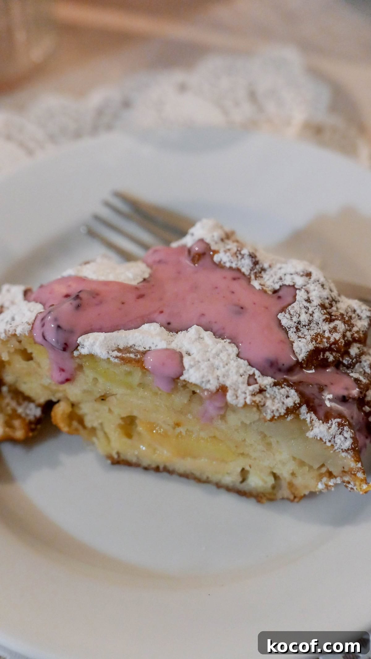 A slice of cake with powdered sugar and pink sauce sits on a white plate, ready to be enjoyed with a fork. This delightful treat brings a touch of Krakow, Poland, to your table.