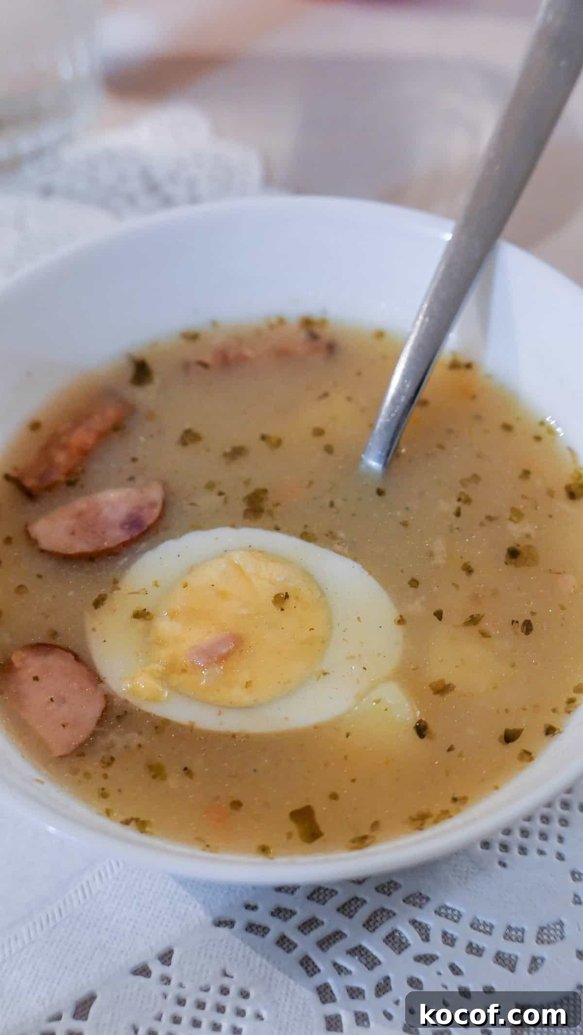 A bowl of soup reminiscent of Krakow, Poland, featuring sliced sausages, fresh herbs, and a halved boiled egg, with a spoon resting inside.