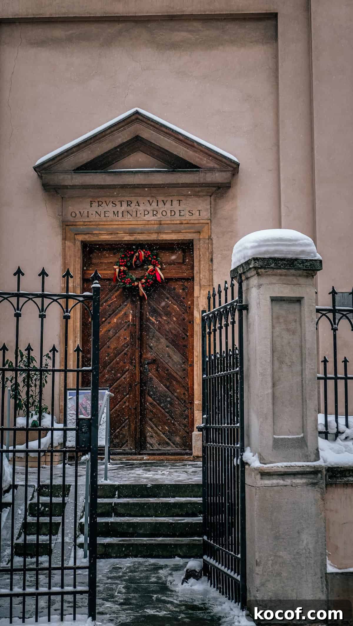 Snow-covered stone building entrance with a wooden door, adorned with a wreath, and a black metal gate in the foreground. A Latin inscription graces the doorway of this charming Krakow, Poland setting.
