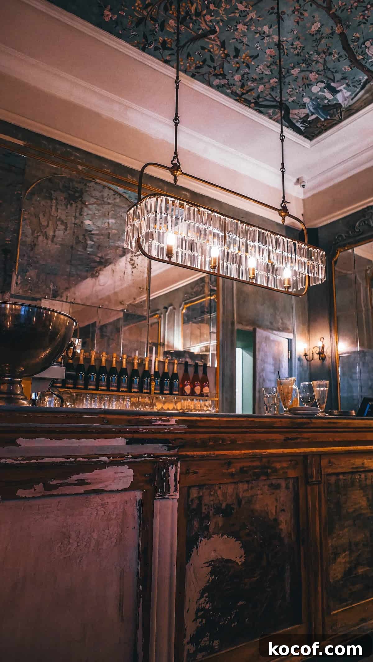 The dimly lit vintage bar in Krakow, Poland features a crystal chandelier, a rustic wooden counter, and bottles lined up against a mirrored wall.