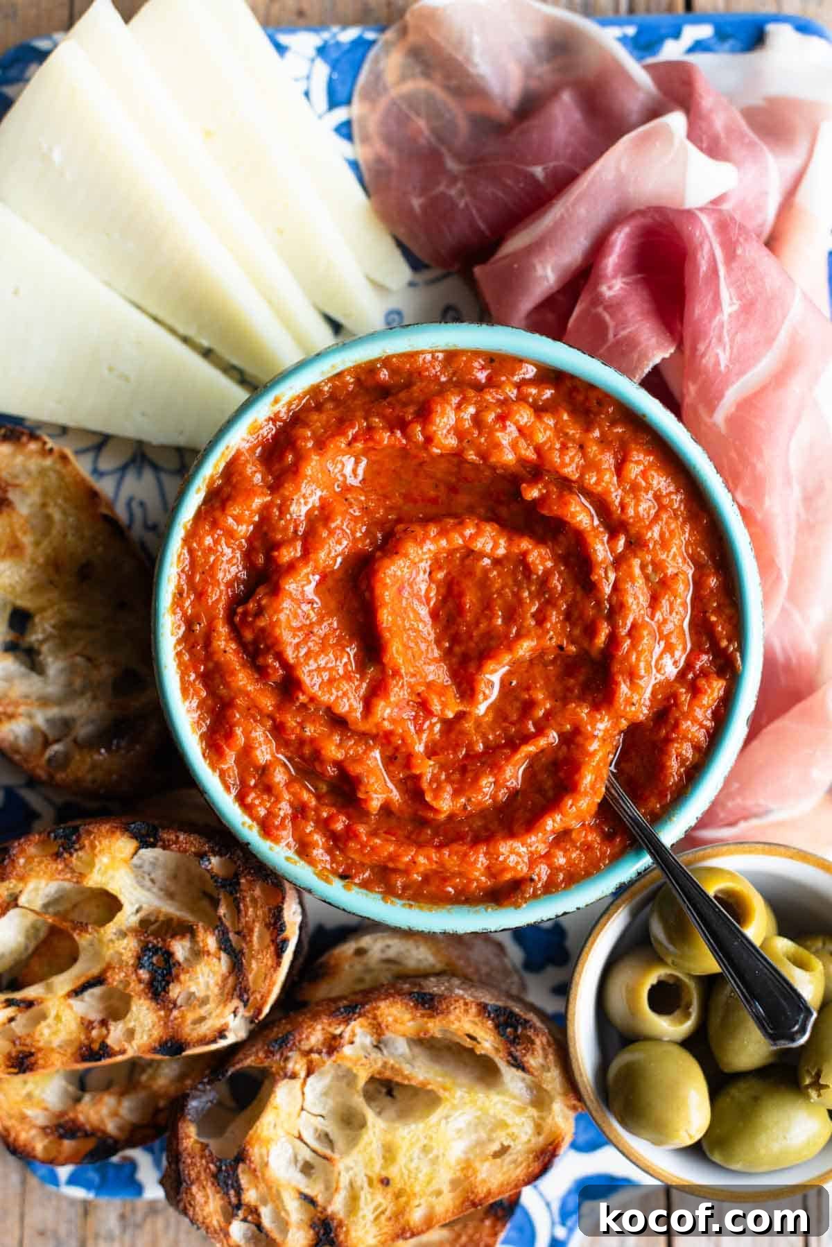 A close up of Ajvar dip in a blue bowl surrounded by cured meat, cheese, ciabatta bread and olives.