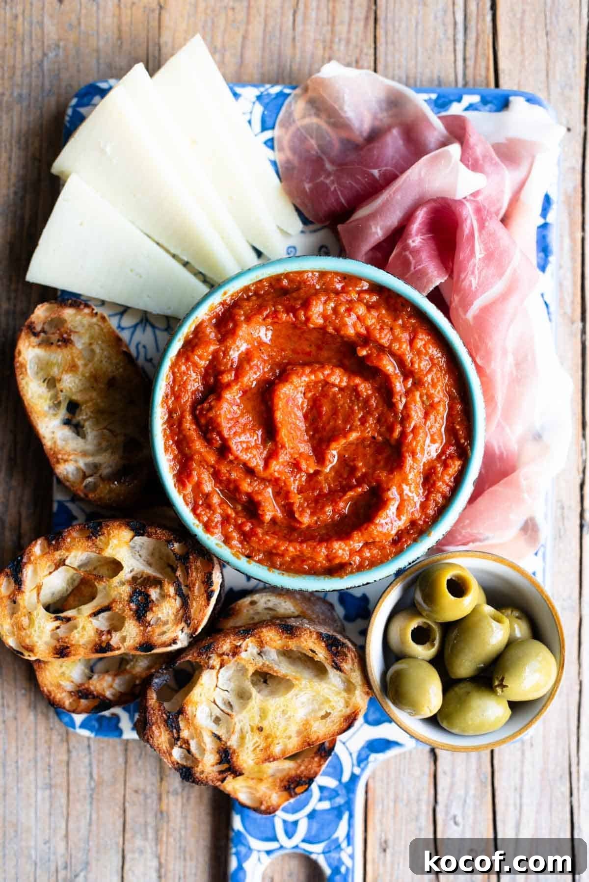An overhead shot of Ajvar spread in a blue bowl sitting on a serving board with cured meats, cheese and olives.