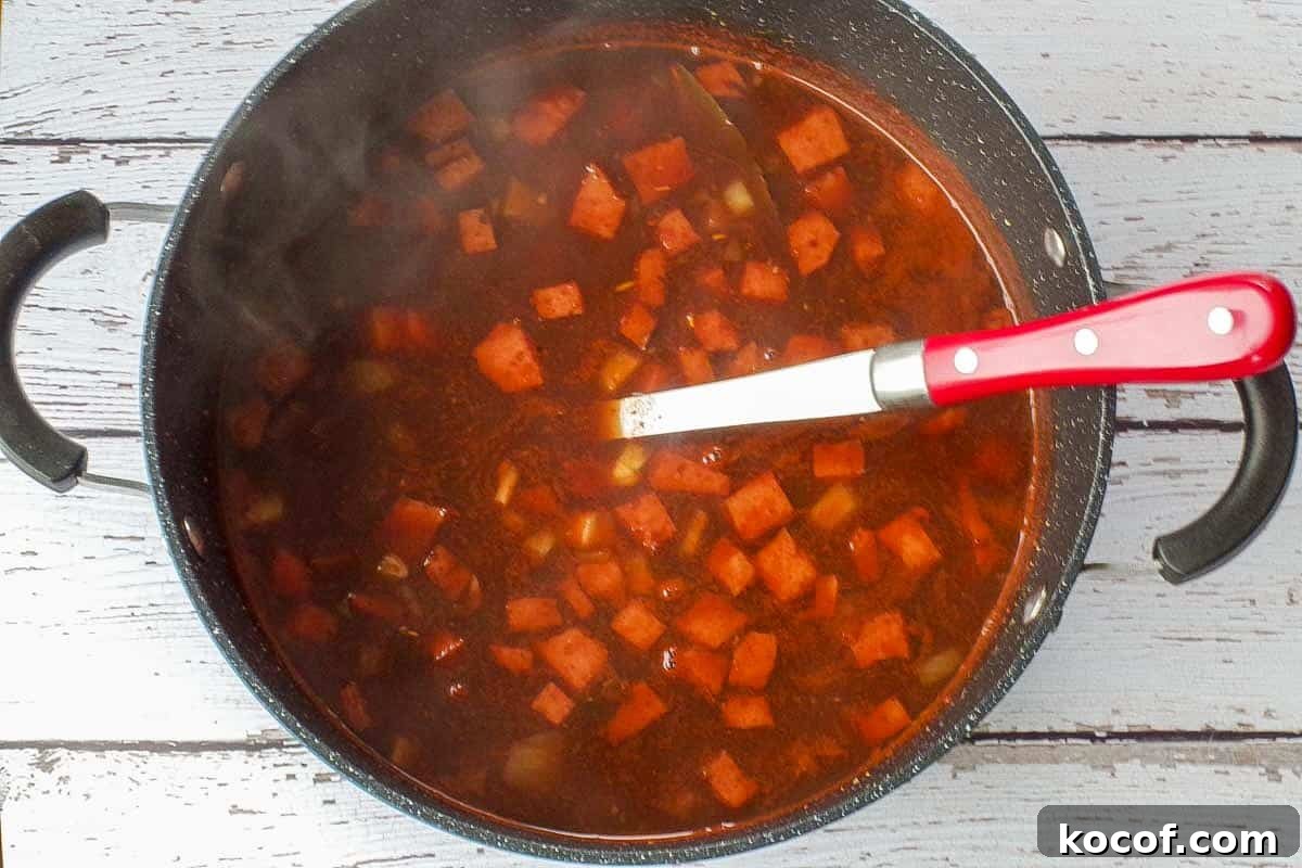Partially cooked ham macaroni soup before adding pasta, in a large pot, with a spoon.