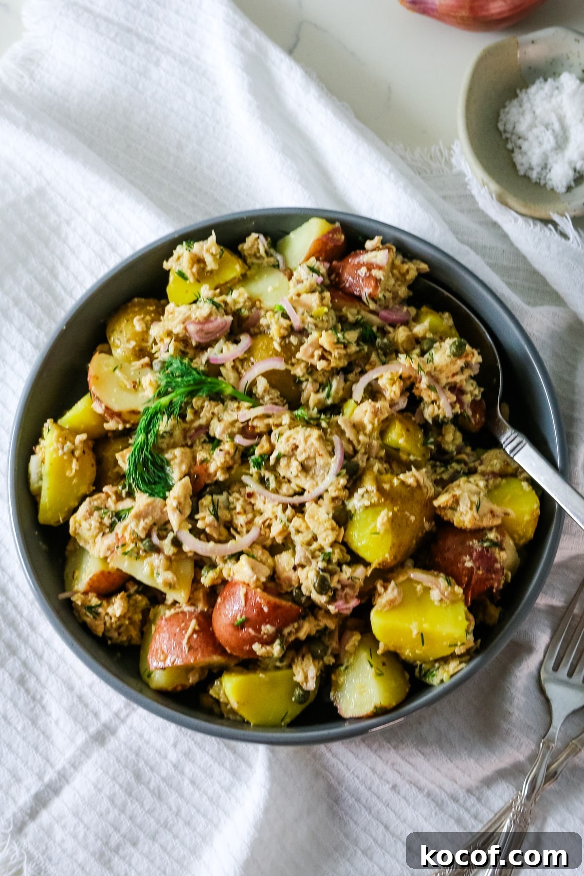 Lemon Dill Tuna & Potato Salad with Light Vinaigrette 6 A gray bowl filled with tuna potato salad with a spoon inside and salt on the side.