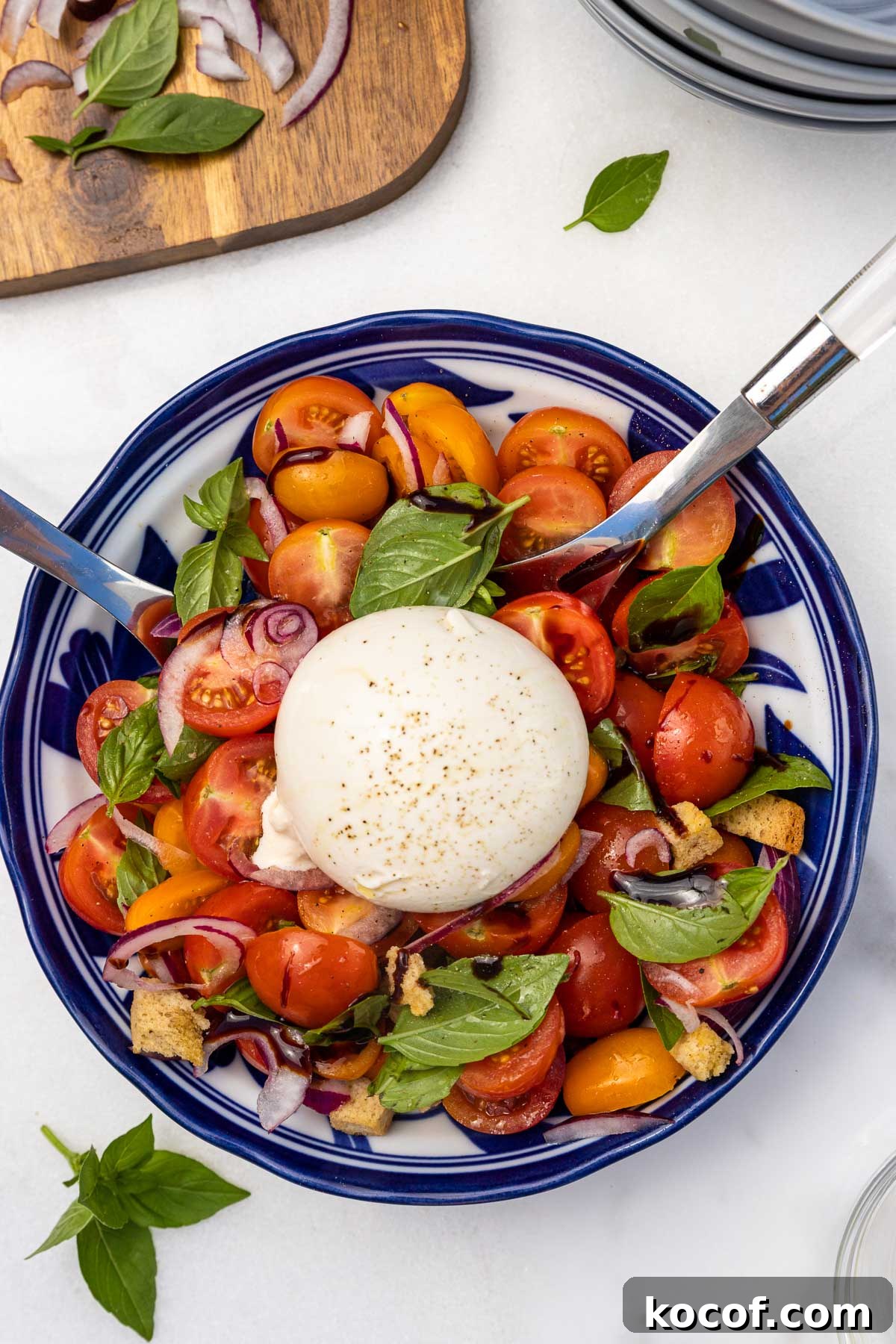 An overhead view of the salad with cherry tomatoes, red onion, croutons and fresh basil in a blue patterned blow with a big whole ball of burrata cheese in the middle, and with serving spoons in it.