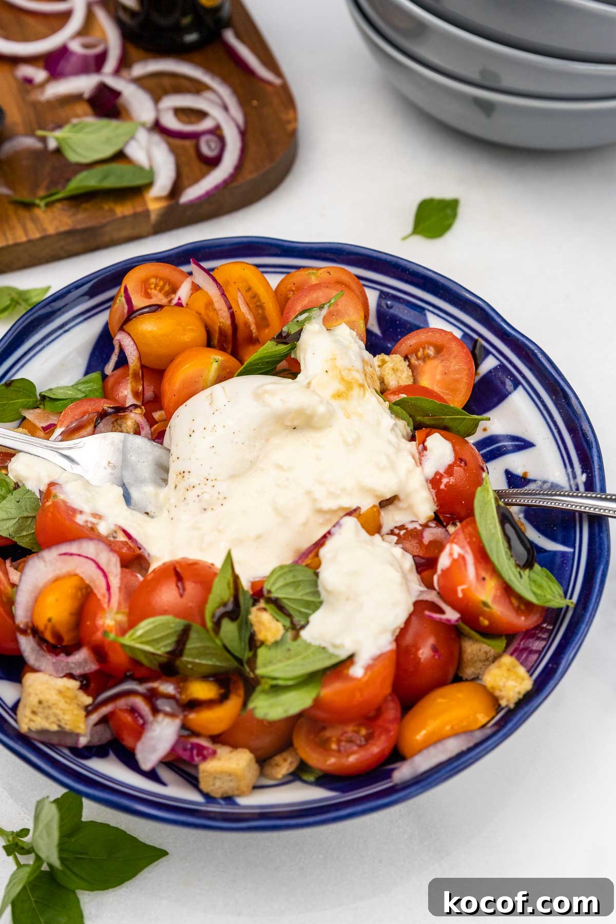 The salad in a blue patterned dish including tomatoes, red onion, basil and croutons with the burrata split open on top, and with a spoon and fork in it.