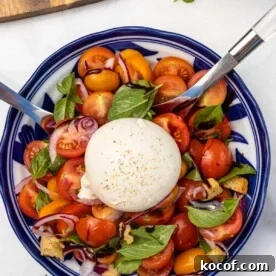 An overhead view of a burrata caprese salad with cherry tomatoes, red onion, croutons and fresh basil in a blue patterned blow with a big whole ball of burrata cheese in the middle, and with serving spoons in it.