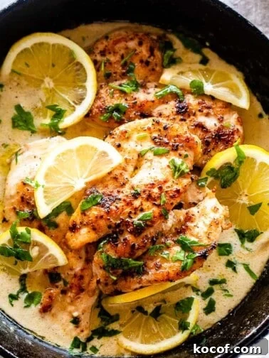 Overhead view of lemon parmesan chicken in a skillet with creamy sauce, topped with lemon slices and fresh herbs.