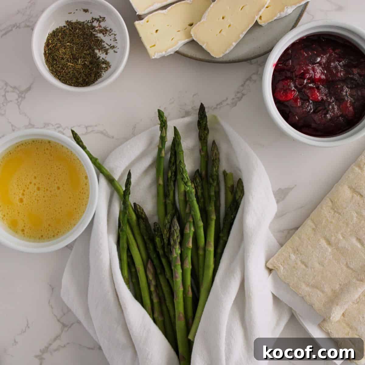 Crispy Puff Pastry-Wrapped Asparagus Recipe 3 Aspargus, egg, rosemary, cranberry jam and brie on a table.