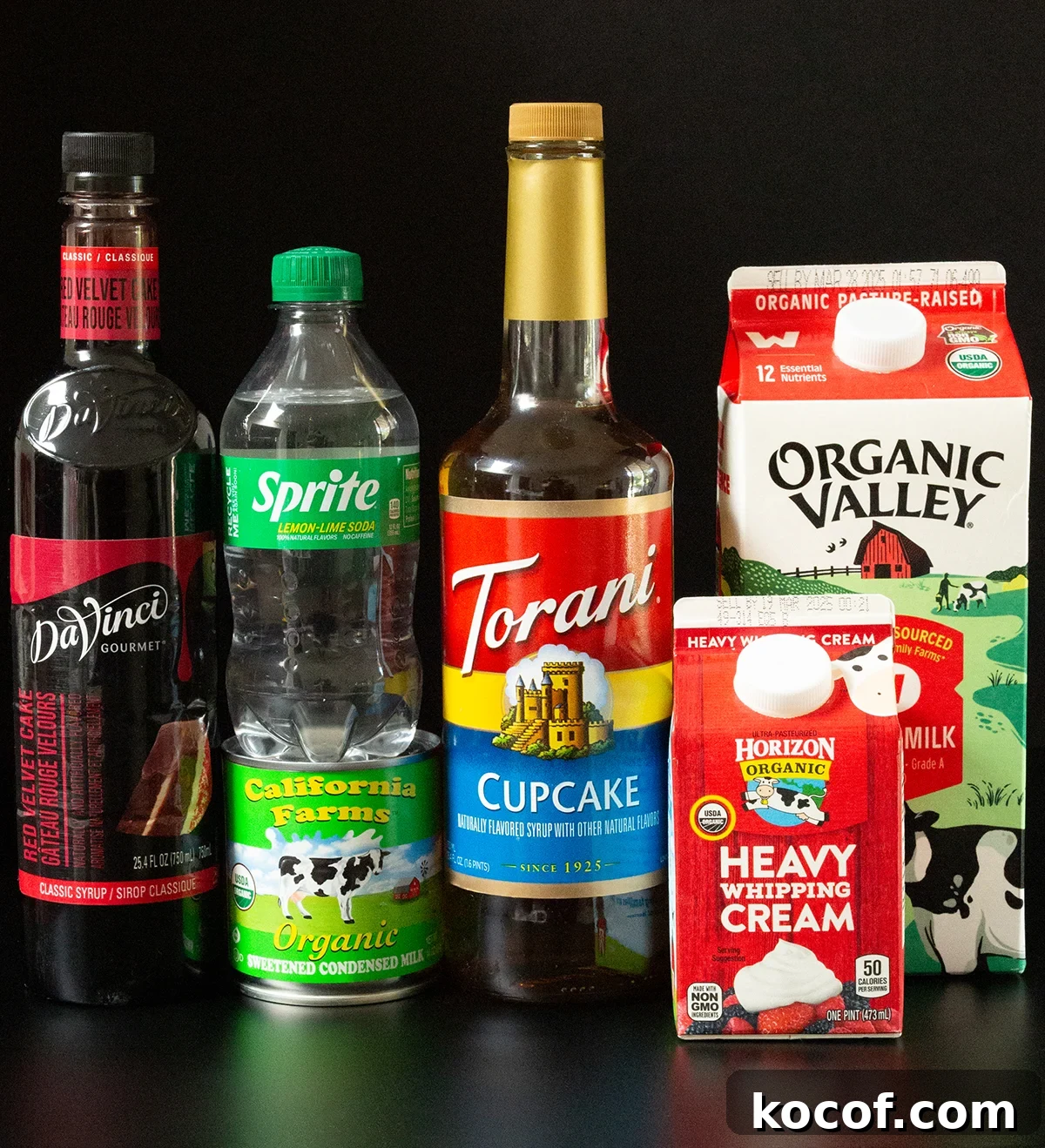 Red Velvet Sprite Cocktail Recipe for Parties and Holidays 3 A bottle of red velvet cake syrup, cupcake syrup, Sprite, a can of sweetened condensed milk, as well as a carton of whole milk and heavy whipping cream all on a black background.