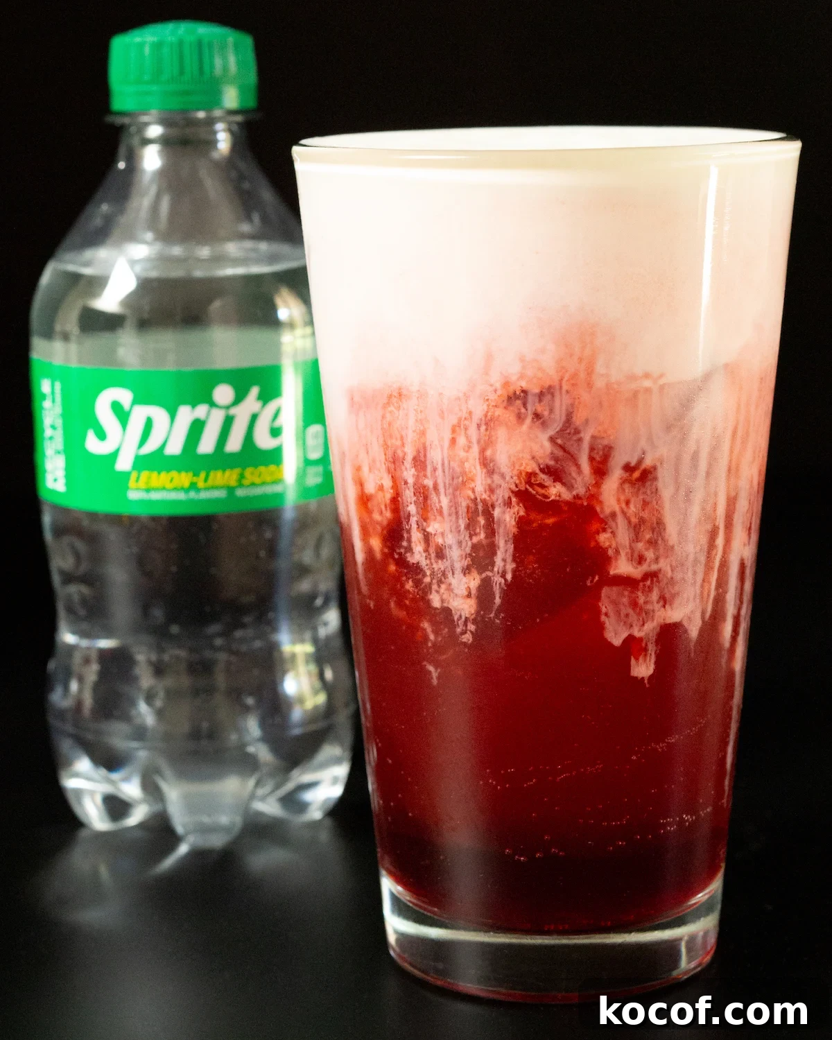 Red Velvet Sprite Cocktail Recipe for Parties and Holidays 4 Most of the frame is taken up by a pint glass filled with a deep red bubbly soda and a thick white cold foam that's starting to bleed into the soda. A Sprite bottle is out of focus in the background.