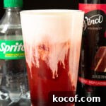 Red Velvet Sprite Cocktail Recipe for Parties and Holidays 5 A pint glass filled with a deep red soda that's topped with a thick white cold foam top. Out of focus in the background is a bottle of sprite and a bottle of red velvet cake syrup.