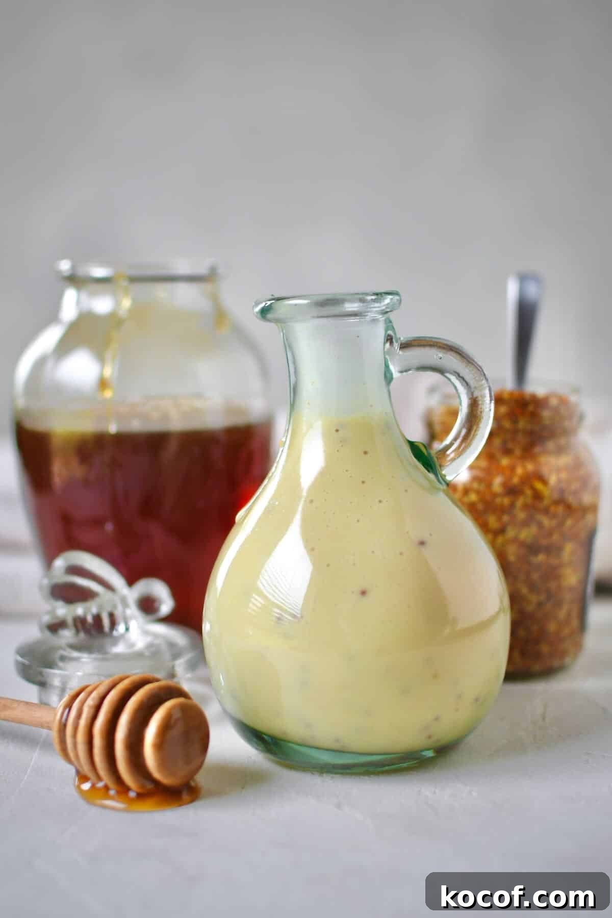 Creamy Honey Mustard Dressing Recipe for Salads & Dipping 2 Dressing mixed up and placed in a storage bottle for later use. A honey jar and mustard jar sit behind it with a dripping honey dipper in front.