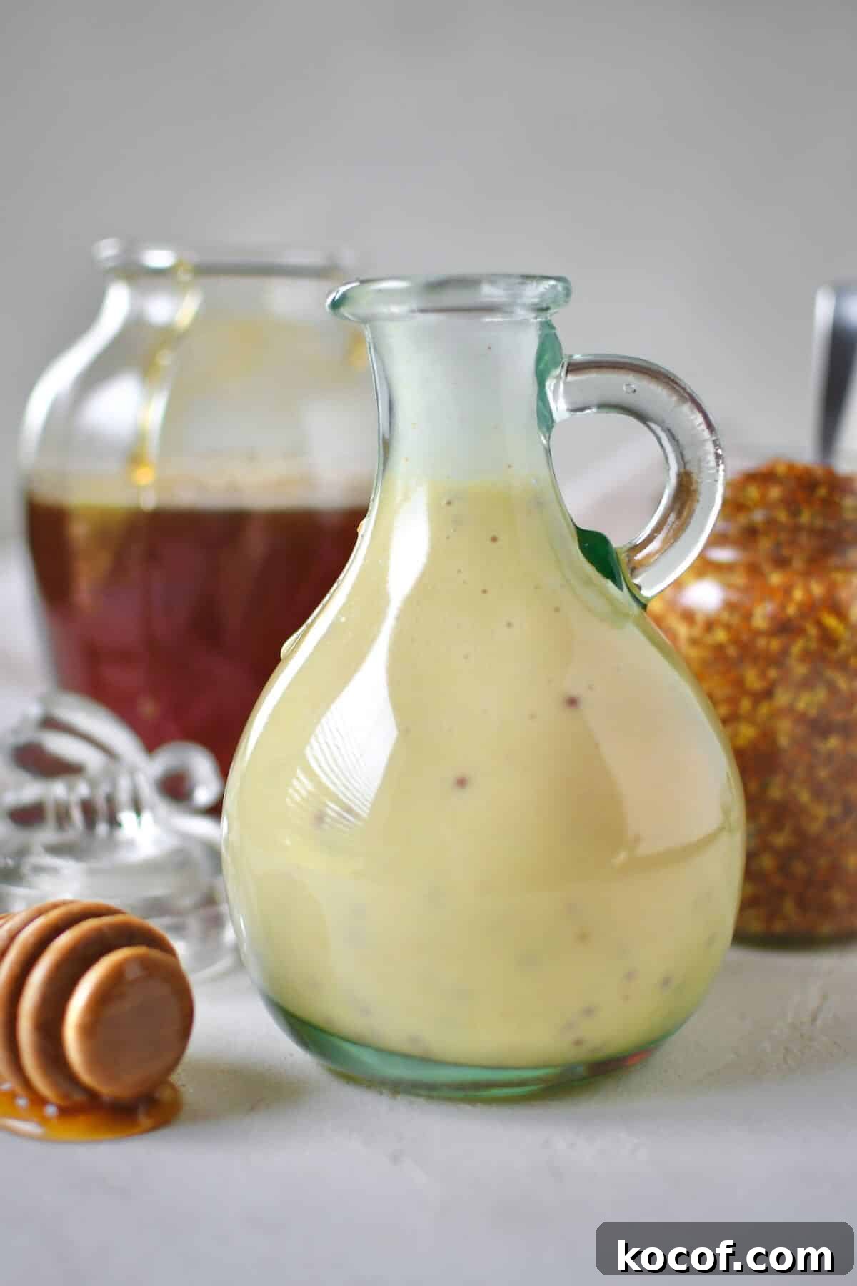 Creamy Honey Mustard Dressing Recipe for Salads & Dipping 7 Dressing mixed up and placed in a storage bottle for later use. A honey jar and mustard jar sit behind it with a dripping honey dipper in front.