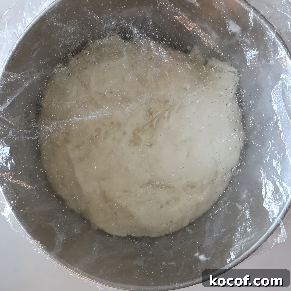 risen dough in stainless steel bowl covered with plastic wrap.
