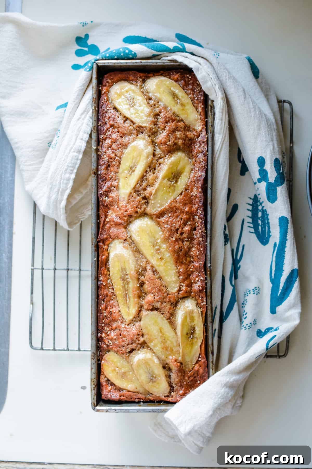 22 Delicious Easter Dinner Ideas That Aren’t Ham 19 A pan of banana bread with sliced bananas on top on a cooling rack with a towel wrapped around the pan.
