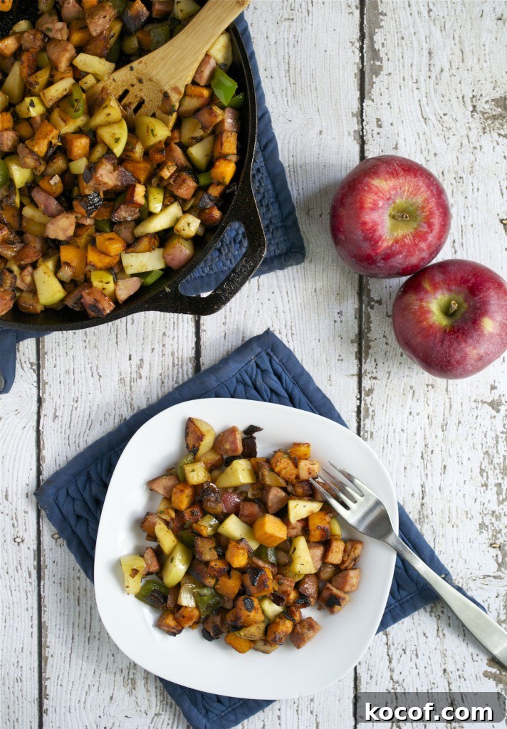 Sweet Potato, Apple & Sausage Hash: Savory Breakfast Skillet 2 A white plate with colorful Apple and Sausage Breakfast Hash—diced apples, sweet potatoes, sausage, and herbs—sits on a blue mat next to a fork. A cast iron skillet with more hash and two red apples rest on a white wooden table.