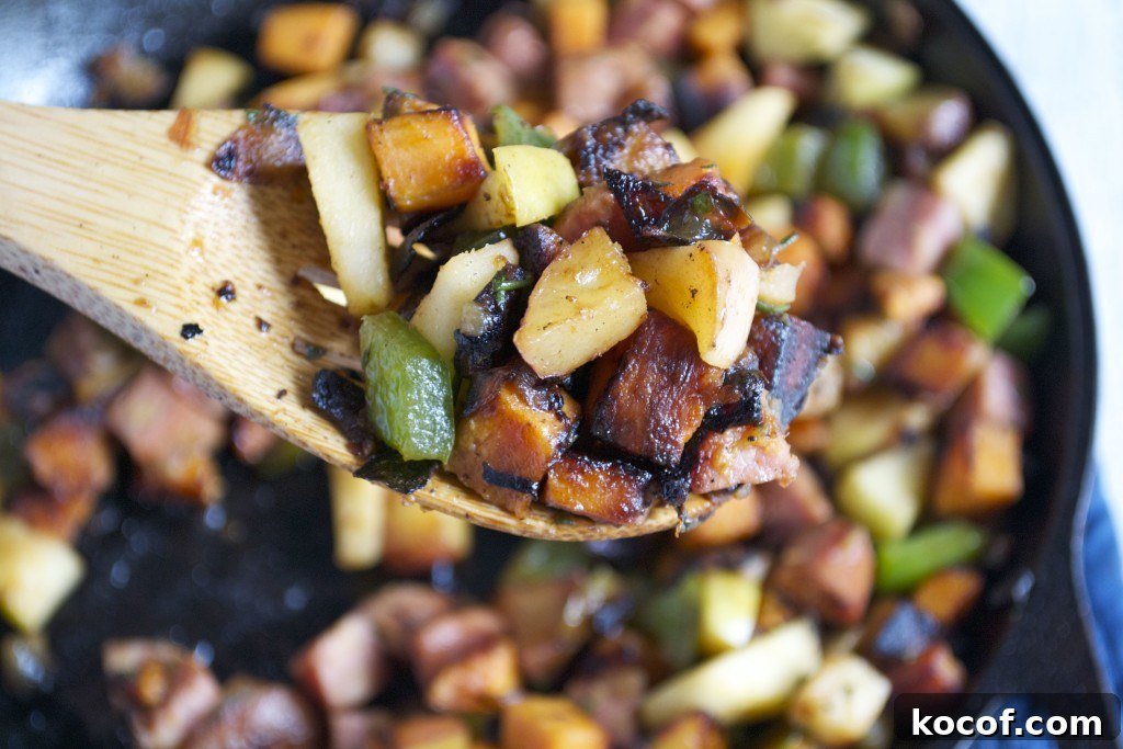 Sweet Potato, Apple & Sausage Hash: Savory Breakfast Skillet 3 A close-up of a wooden spoon lifting a mix of sautéed potatoes, sweet potato, green bell peppers, onions, and sausage from a skillet. The vegetables and sausage are browned and cooked for an Apple and Sausage Breakfast Hash.