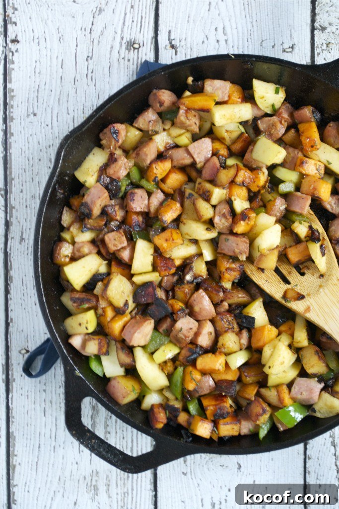 Sweet Potato, Apple & Sausage Hash: Savory Breakfast Skillet 4 A cast iron skillet filled with Sweet Potato, sausage, green peppers, and onions sits on a white wooden surface. A wooden spatula rests in the skillet, mixing the browned and colorful ingredients for a delicious Apple and Sausage Breakfast Hash.